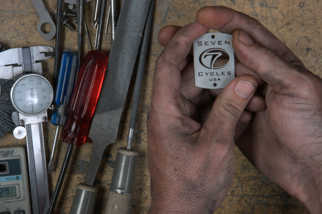 Working hands hold a Seven headbadge near some tools on a workbench