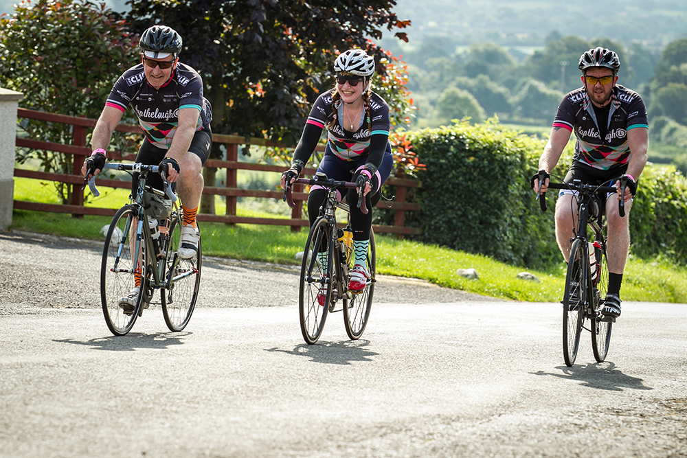 Three cyclists smile and laugh together as they ride up a country road on a sunny summer day