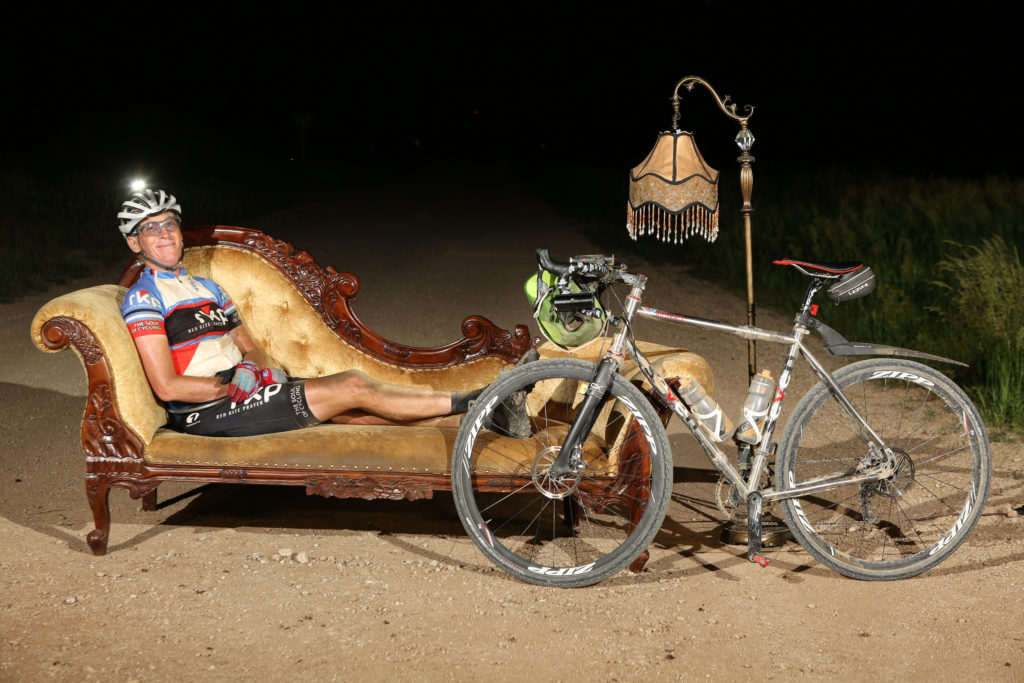 Patrick Brady in his cycling kit reclines on a chaise lounge with an antique lamp on a night road. His bike is propped by his side.